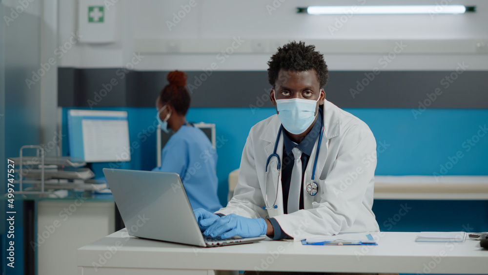 Portrait of young specialist using laptop sitting at desk while wearing gloves and face mask in medical office. Doctor typing on device and nurse with computer in background during pandemic