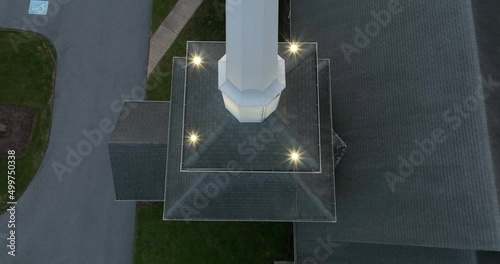 Tall white steeple on church at night, lights on. Rising aerial reveals weather vane. Top down view. photo