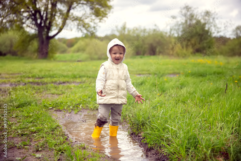 Cute child toddler in a warm vest and jacket stands in yellow rubber boots in a large puddle, Free children's walks in nature, mud and puddles Cute child toddler in a warm vest and jacket stands in yellow rubber boots in a large puddle, Free children's walks in nature, mud and puddles