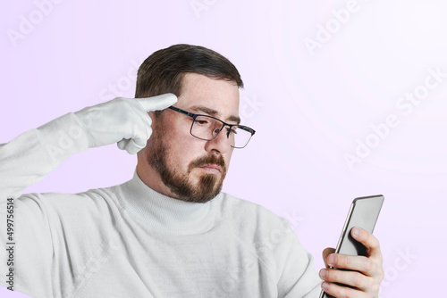 Portrait of an attractive bearded guy in glasses pointing a finger in a white glove at his head. The man is holding a phone. Isolated pastel colored background.