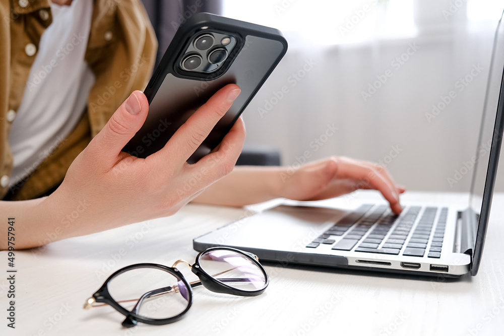 Fototapeta premium Close up of female hands holding smartphone and typing on computer keyboard, designer or student using laptop sitting on sofa at home, online learning, internet marketing, office workplace concept