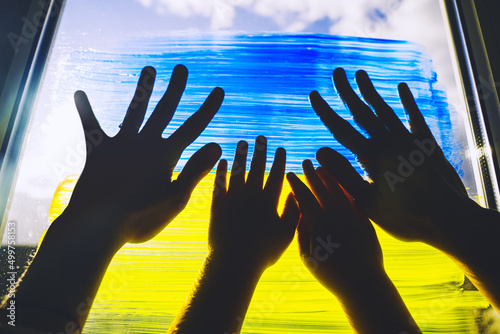 Human hands of parent and child touch painting yellow-blue flag of Ukraine on window. Adult and kids hands on image of flag of Ukraine on glass. Support Ukrainians. Concepts symbol.