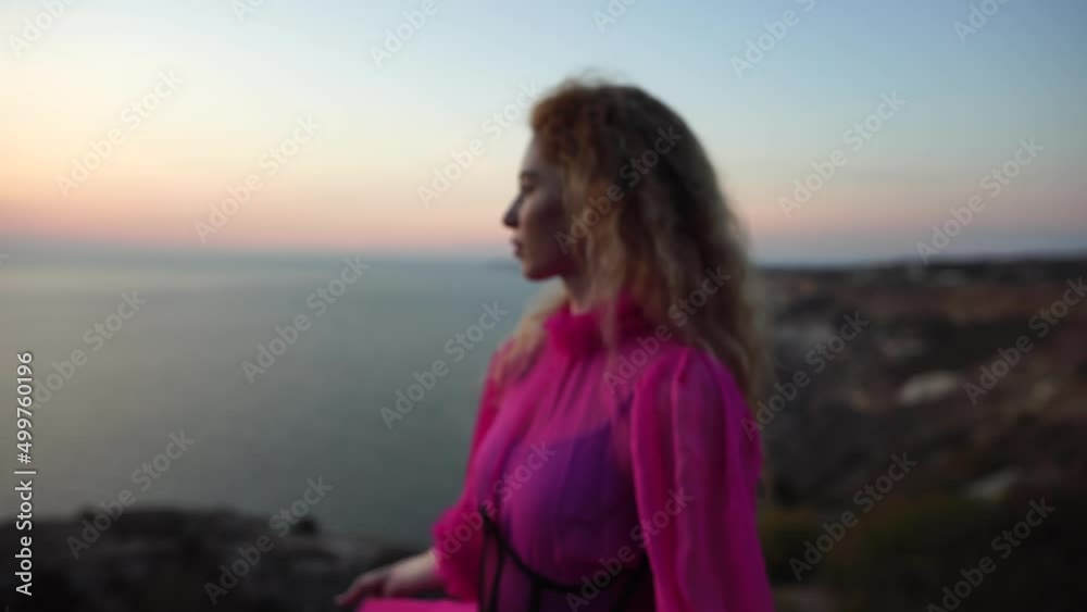 Abstract defocused shot of beautiful young caucasian woman with curly blond hair and freckles looking at camera. Cute woman portrait in a pink long dress posing on a volcanic rock high above the sea Abstract defocused shot of beautiful young caucasian woman with curly blond hair and freckles looking at camera. Cute woman portrait in a pink long dress posing on a volcanic rock high above the sea