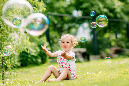 A little girl catches soap bubbles in the park, sitting on the grass. The concept of a happy childhood