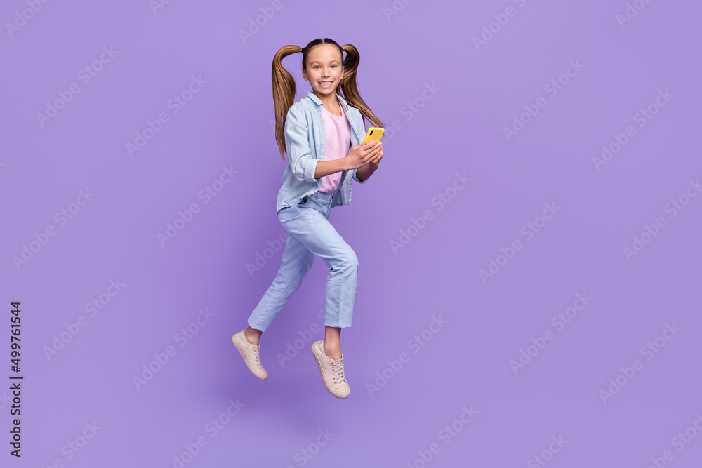 Photo of shiny adorable little school girl wear denim shirt jumping typing modern device empty space isolated purple color background