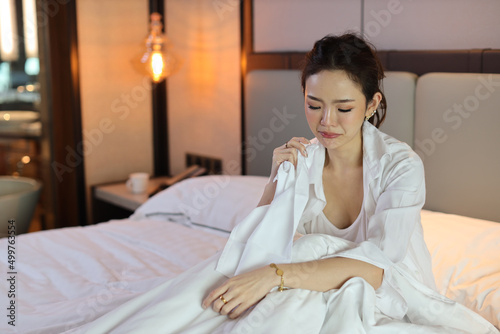 Young depressed asian woman sitting and holding handkerchief while crying on bed in hotel bedroom with low light environment. Sad girl, unhappy and loneliness concept.