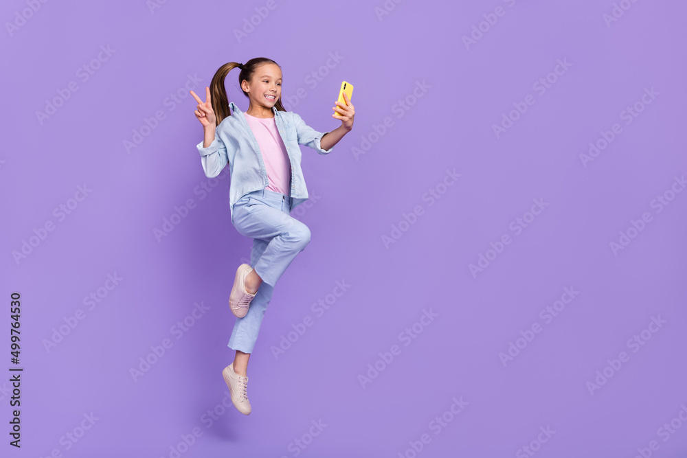 Photo of cute shiny little school girl wear denim shirt jumping tacking selfie device v-sign empty space isolated purple color background Photo of cute shiny little school girl wear denim shirt jumping tacking selfie device v-sign empty space isolated purple color background