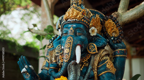 An old stone statue of Ganesha covered with moss with a wreath of yellow flowers on his neck standing on the street with green plants