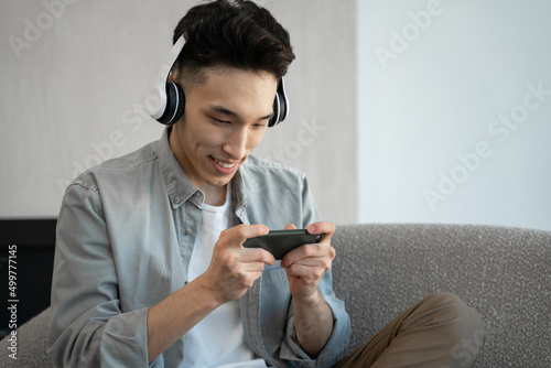 Focused Asian man in headphones enjoys playing racing game on smartphone. Brunet guy sits on soft couch in living room having fun at home closeup