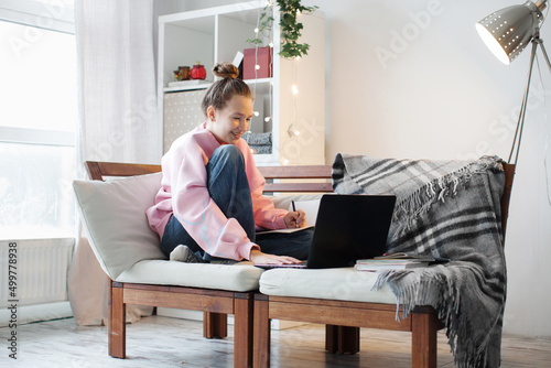 Millennial teen girl sit at couch in living room study on laptop making notes