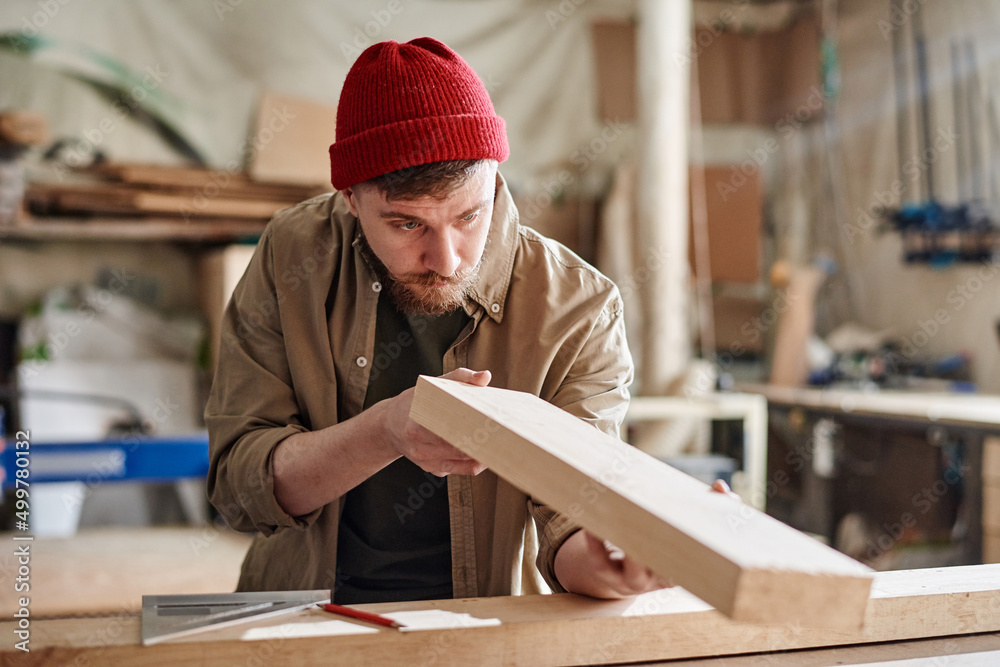 Horizontal medium portrait of young Caucasian man working in joinery workshop checking wooden plank surface before polishing it