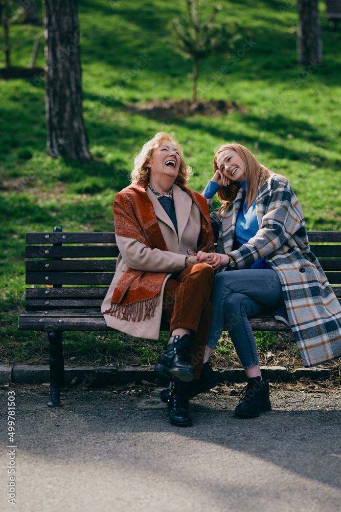 A grandmother is laughing and having fun with her adolescent granddaughter while sitting on the park bench. A grandmother is laughing and having fun with her adolescent granddaughter while sitting on the park bench.
