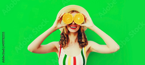 Summer portrait of happy cheerful smiling woman covering her eyes with slices of orange and looking for something wearing straw hat on green background