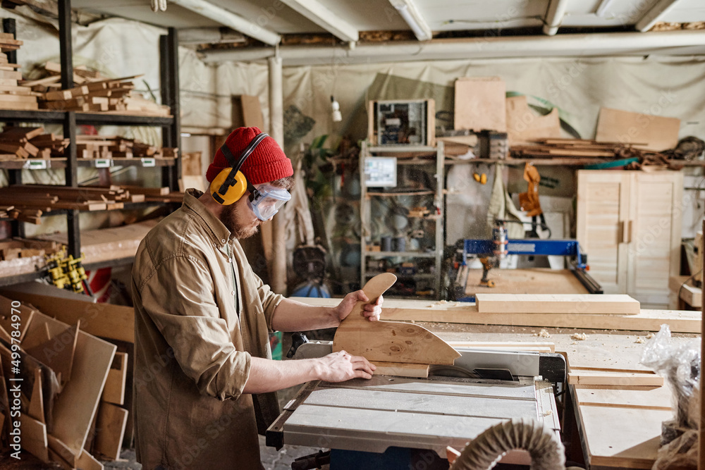 Young male carpenter wearing safety glasses and hearing protection headphones cutting wood plank with table saw
