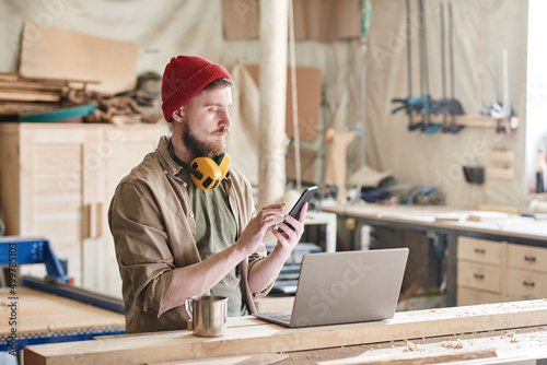 Valokuva Modern woodworker standing at workbench with laptop and cup of tea on it texting