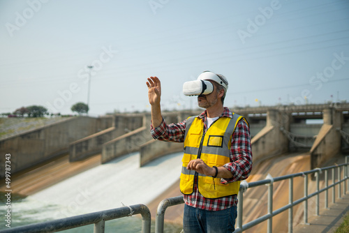 Portrait of engineer wearing yellow vest and white helmet using augmented reality application in virtual reality glasses. Working on a dam with a hydroelectric power plant. Sustainable energy concept