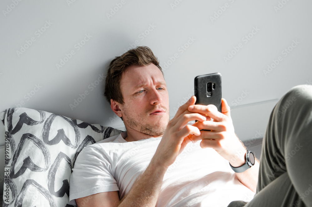 Caucasian attractive young man in white t-shirt woke up chatting, surfing, working on pc and making phone call on bed in bedroom interior. Modern device for remote communication Caucasian attractive young man in white t-shirt woke up chatting, surfing, working on pc and making phone call on bed in bedroom interior. Modern device for remote communication