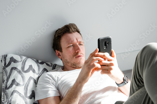 Caucasian attractive young man in white t-shirt woke up chatting, surfing, working on pc and making phone call on bed in bedroom interior. Modern device for remote communication