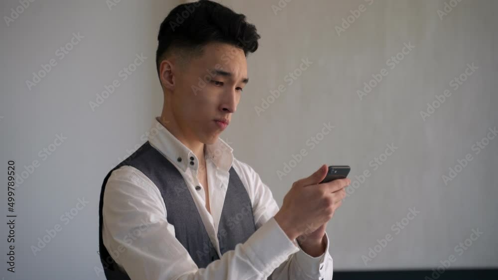 Young Asian businessman in formal clothes chooses outfit for party in online store. Picky man looks in smartphone shaking head in room closeup Young Asian businessman in formal clothes chooses outfit for party in online store. Picky man looks in smartphone shaking head in room closeup