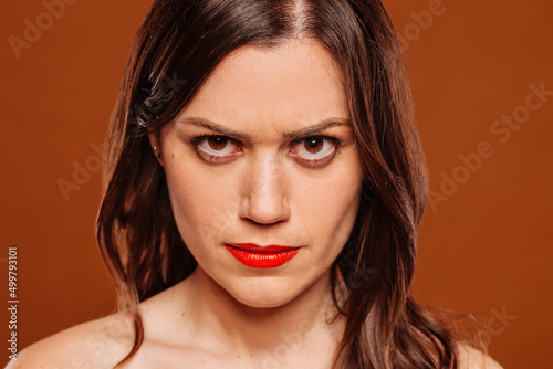 Obraz na plátně Face portrait of young angry hateful woman looking at camera at studio over brown background
