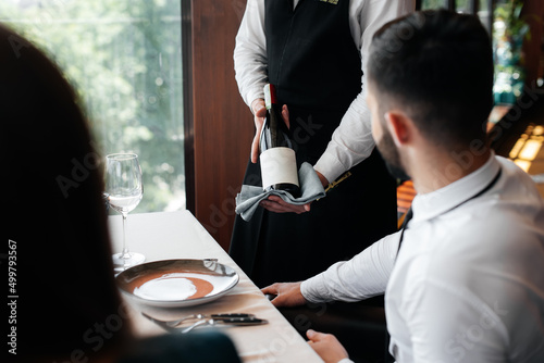 A young sommelier in a stylish apron demonstrates and offers fine wine to a beautiful couple in a restaurant. Customer service in an elite restaurant and a public restaurant. photo