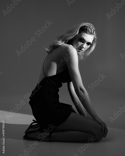 Young sexy fashionable blonde woman with long legs in designer 2022 trend evening short black silk dress. Vogue beauty glamour style. Studio shot. Creative light
