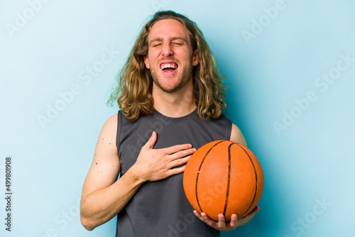 Young caucasian man playing basketball isolated on blue background laughs out loudly keeping hand on chest.