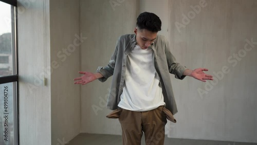 Asian man looks for money putting hands into inside-out pockets. Poor guy in casual clothes feels disappointed standing in empty room near big window photo