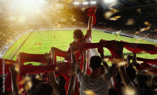 Rear view of football, soccer fans cheering their team with scarfs at crowded stadium at evening time. Concept of sport, support, competition. Out of focus effect