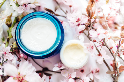 Top view of cosmetic cream and concealer eyes creams with pink cherry flowers in a blue glass jar. Hygienic skincare lotion product.