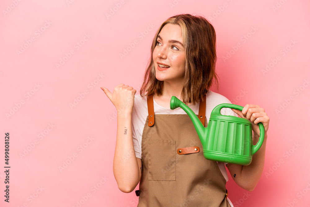 Young gardener woman holding watering can isolated on pink background points with thumb finger away, laughing and carefree. Young gardener woman holding watering can isolated on pink background points with thumb finger away, laughing and carefree.