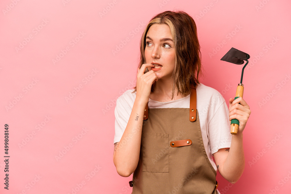 Young gardener woman holding gardening shovel isolated on pink background relaxed thinking about something looking at a copy space.