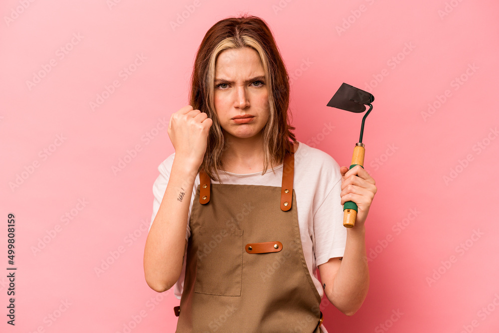 Young gardener woman holding gardening shovel isolated on pink background showing fist to camera, aggressive facial expression. Young gardener woman holding gardening shovel isolated on pink background showing fist to camera, aggressive facial expression.