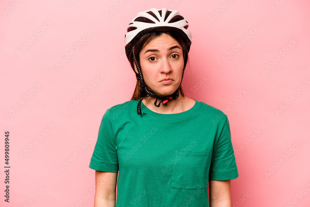 Young caucasian woman wearing helmet isolated on pink background shrugs shoulders and open eyes confused.