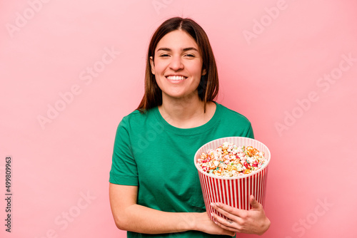 Young caucasian woman holding popcorn isolated on pink background laughing and having fun.