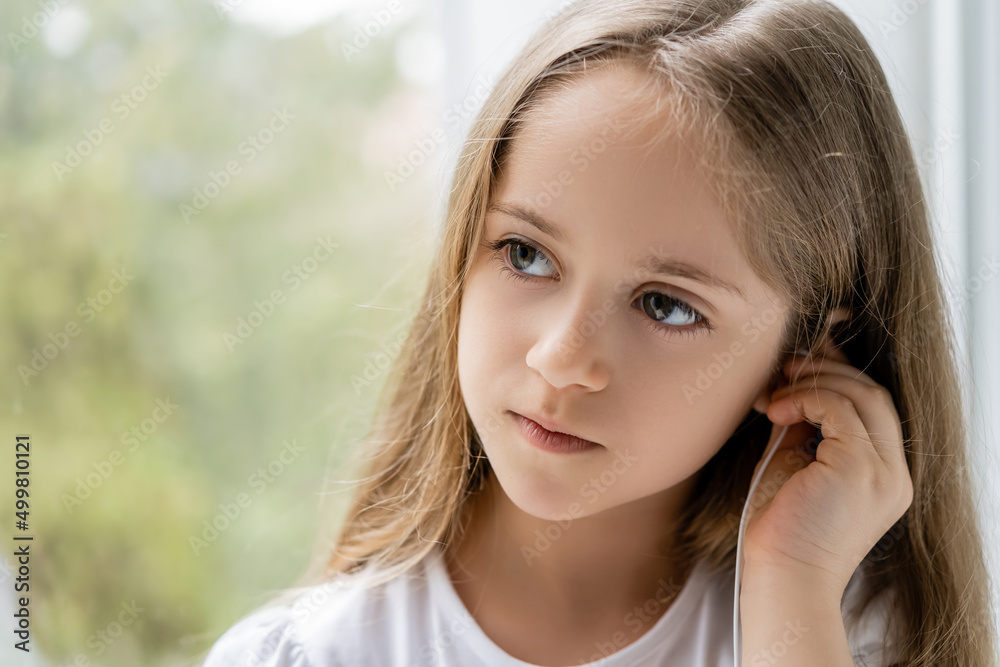 girl with long hair adjusting earphone near blurred window.