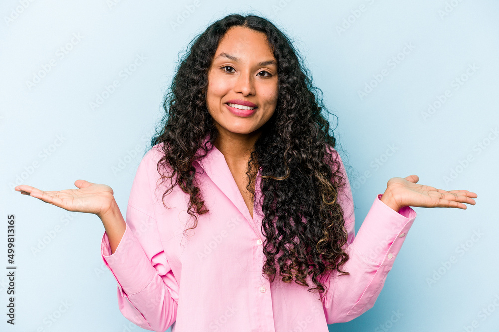 Young hispanic woman isolated on blue background makes scale with arms, feels happy and confident. Young hispanic woman isolated on blue background makes scale with arms, feels happy and confident.