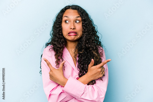 Young hispanic woman isolated on blue background points sideways, is trying to choose between two options.