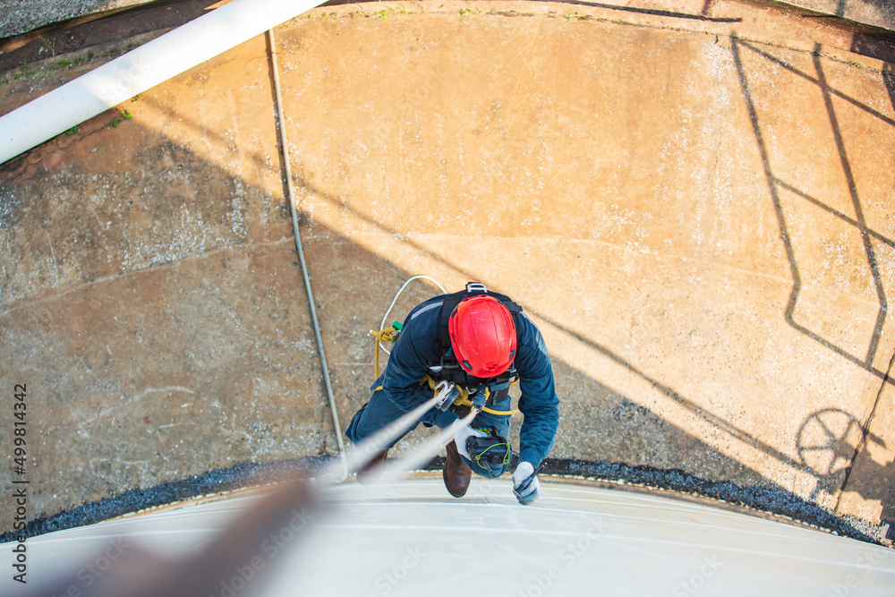 Top view pic of industrial rope access welder working at height wearing harness, helmet safety equipment rope access inspection of thickness storage tank Top view pic of industrial rope access welder working at height wearing harness, helmet safety equipment rope access inspection of thickness storage tank