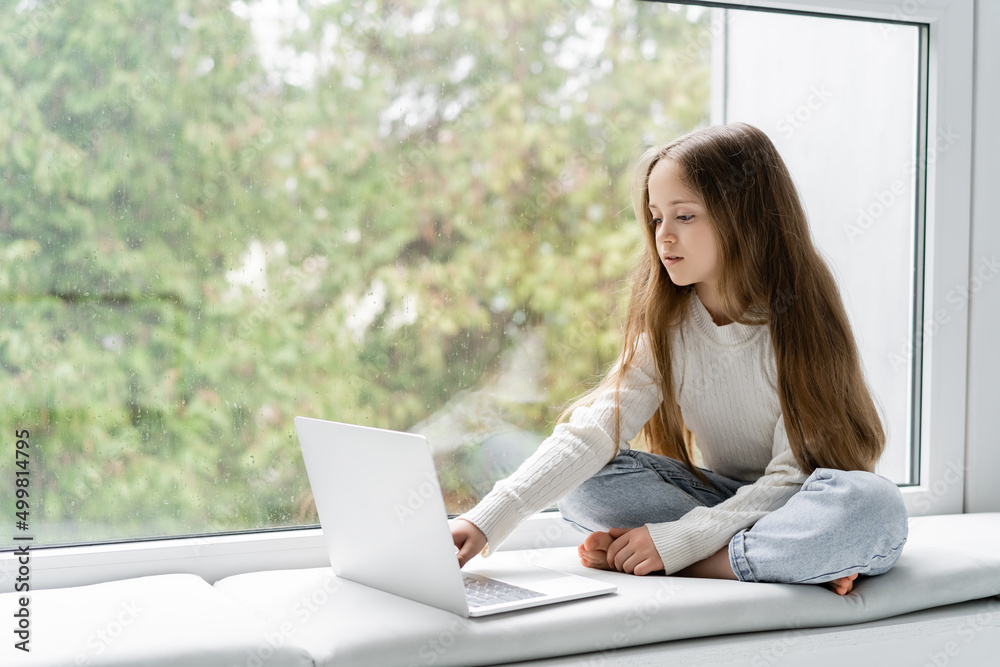 girl sitting on windowsill with crossed legs and using laptop during online lesson. girl sitting on windowsill with crossed legs and using laptop during online lesson.