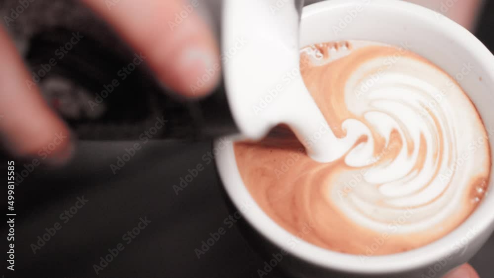 barista hands making latte art cappuccino pouring frothed milk from pitcher into white ceramic mug with espresso coffee in ultra slow motion closeup
