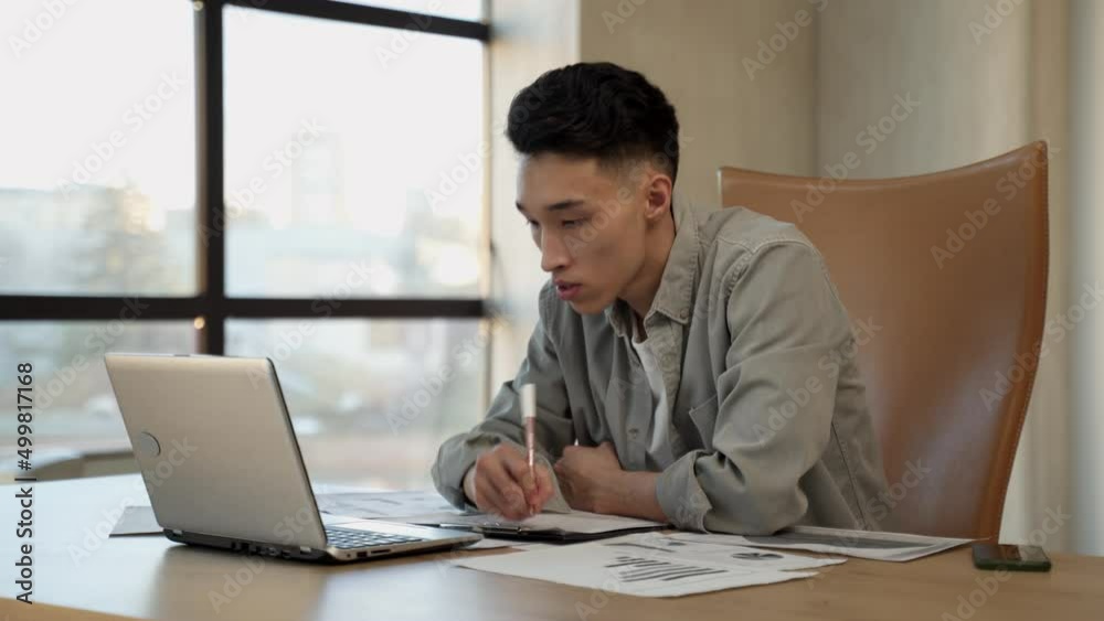 Young Asian guy manager gets happy for successful project writing down details. Man works near laptop at table sitting in office near bright window Young Asian guy manager gets happy for successful project writing down details. Man works near laptop at table sitting in office near bright window