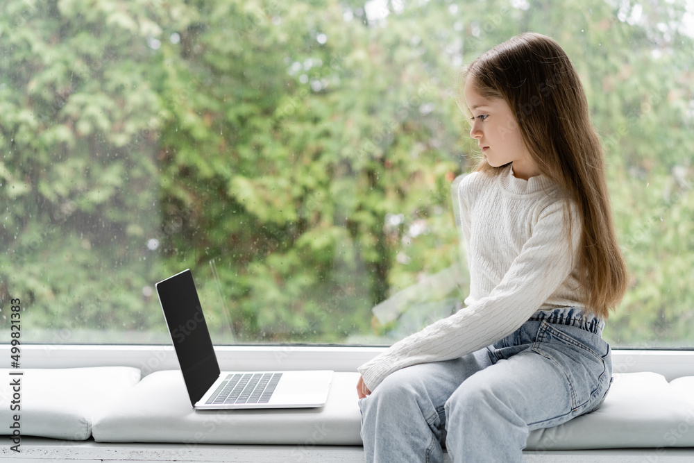 girl sitting on windowsill at home near laptop with blank screen. girl sitting on windowsill at home near laptop with blank screen.