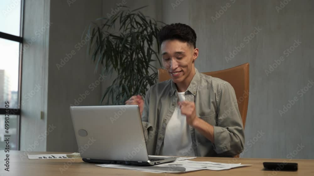 Cheerful Asian employee has fun at end of working day in office. Young employee moves on chair near laptop at wooden table and lush flower-plant Cheerful Asian employee has fun at end of working day in office. Young employee moves on chair near laptop at wooden table and lush flower-plant