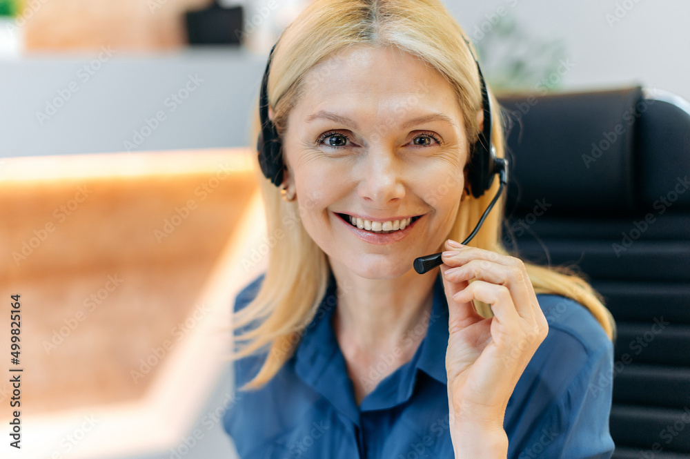 Close-up of positive caucasian attractive confident adult blonde woman with headset, sitting in office, working as operator of call center or support service, looking at the camera, smiling friendly Close-up of positive caucasian attractive confident adult blonde woman with headset, sitting in office, working as operator of call center or support service, looking at the camera, smiling friendly