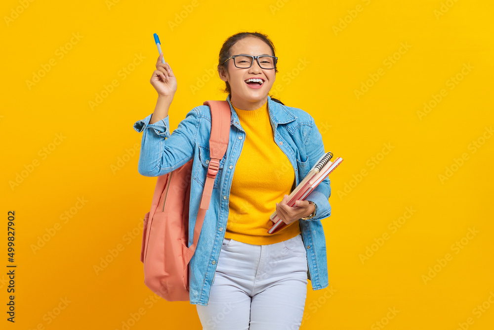Portrait of cheerful young Asian woman student in casual clothes with backpack holding book and pointing at copy space with pen isolated on yellow background. Education in college university concept Portrait of cheerful young Asian woman student in casual clothes with backpack holding book and pointing at copy space with pen isolated on yellow background. Education in college university concept