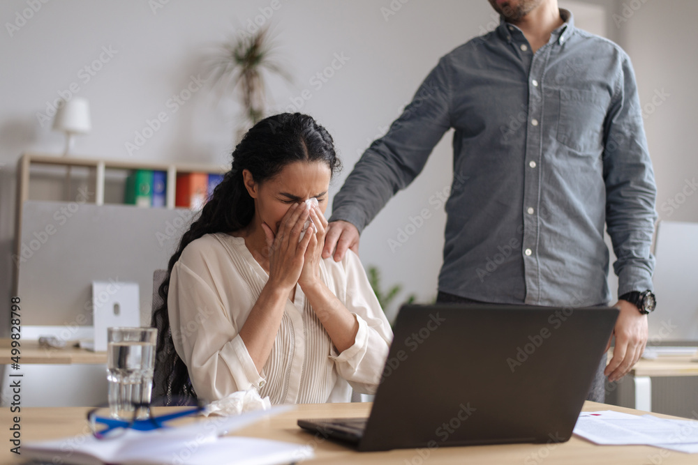 Fototapeta premium Upset young woman crying near laptop pc at office, male colleague helping and comforting her