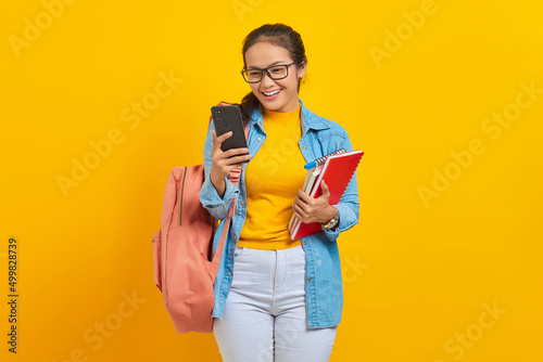 Portrait of cheerful young Asian woman student in denim clothes, glasses with backpack holding mobile phone and books isolated on yellow background. Education in high school university college concept