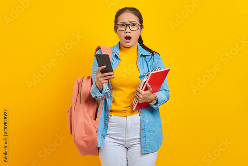 Portrait of amazed young Asian woman student in denim clothes with backpack holding mobile phone and books isolated on yellow background
