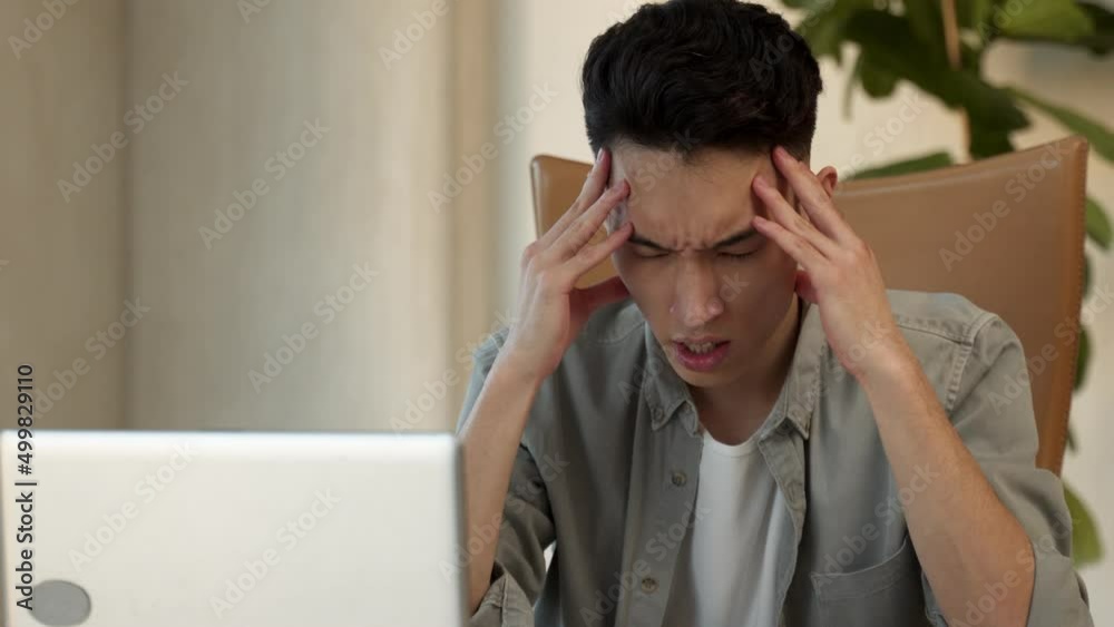 Young Asian male employee holds head trying to solve difficult issue at work in office. Desperate man sits near laptop at table feeling unsure closeup Young Asian male employee holds head trying to solve difficult issue at work in office. Desperate man sits near laptop at table feeling unsure closeup